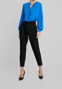 Blå blus med knäppning vid halsen, långärmad; matchad med högmidjade svarta byxor med knytbälte. Svarta spetsiga klackar.