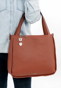 Brun læderhåndtaske med en tekstureret finish, dobbelte håndtag og en sølv charme med mærkets logo. Solidt, struktureret design.