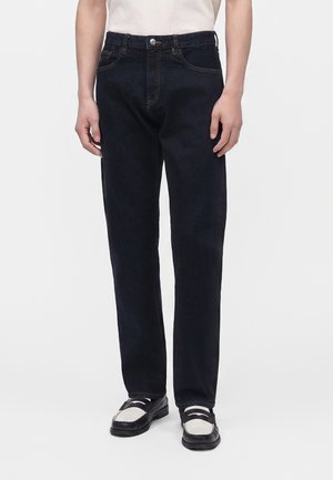 Person trägt dunkelblaue Straight-Leg-Jeans und schwarz-weiße Loafer, steht vor einem schlichten hellen Hintergrund.