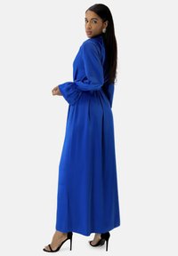 Lungo vestito blu in tessuto liscio con maniche a campana e collo alto. Abbinato a scarpe nere con tacco alto.