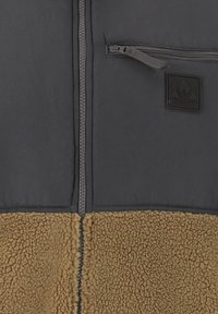Fleecejacka med en svart övre del och en brun shearling-nedre panel. Har en dragkedja framtill och en liten dragkedjeförsedd bröstficka.