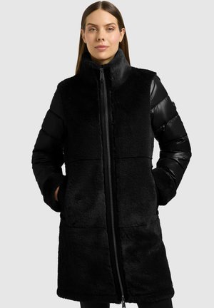 Femme portant un long manteau noir avec devant en polaire et manches matelassées brillantes, debout les mains dans les poches sur un fond clair.