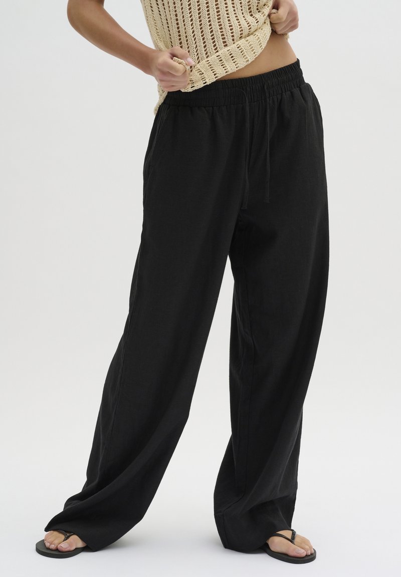 Personne portant un pantalon ample noir avec cordon, un haut sans manches beige en maille et des tongs noires, debout devant un fond blanc uni.