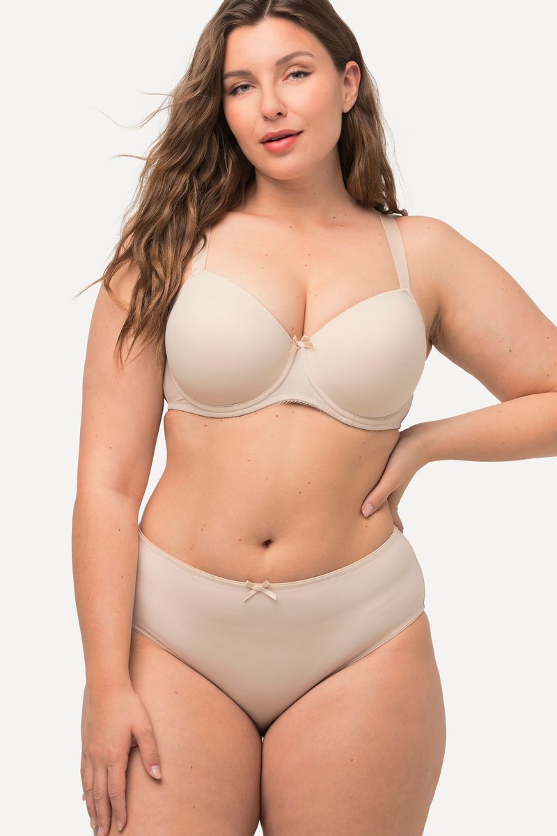Kobieta w rozmiarze plus size z długimi falowanymi włosami, ubrana w beżowy, dopasowany biustonosz i majtki z wysokim stanem, pozująca z jedną ręką na biodrze.
