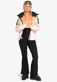Ski-outfit met een zwarte skijas met oranje en crème accenten, een oranje gestreepte tussenlaag, zwarte broek en witte handschoenen.