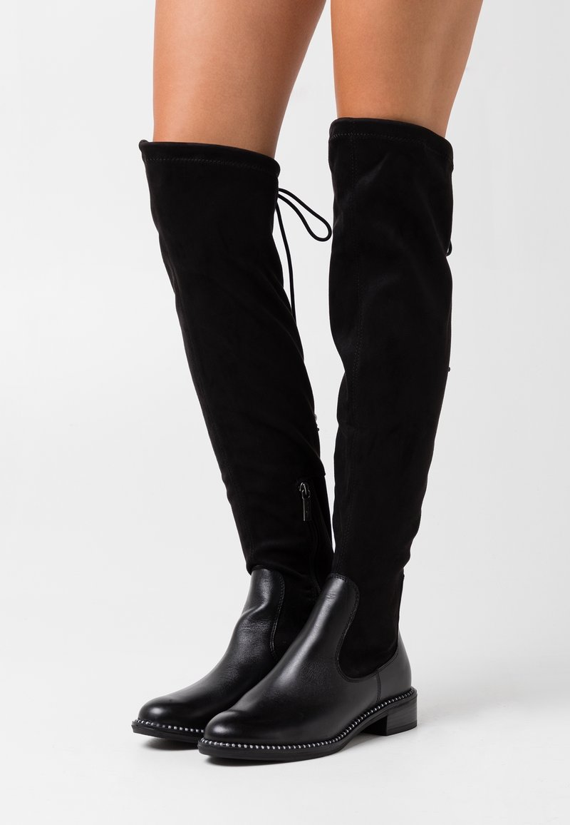 Bottes noires cuissardes avec pied en cuir, tige en daim, fermeture éclair latérale et clous argentés décoratifs le long du bord de la semelle sur fond blanc.
