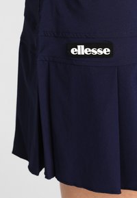 Falda atlética de color azul marino fabricada con tejido ligero y con pliegues; presenta una etiqueta de logo en blanco y negro en la parte frontal.