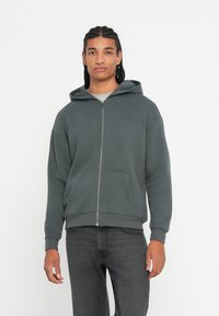 Giovane con i capelli intrecciati, indossa una felpa grigia scura con zip e jeans neri, in piedi su uno sfondo bianco semplice.