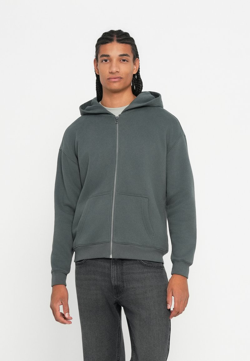 Giovane con i capelli intrecciati, indossa una felpa grigia scura con zip e jeans neri, in piedi su uno sfondo bianco semplice.