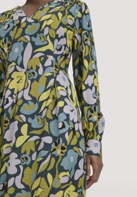 Langärmliges Kleid mit grünem, blauem und lavendelfarbenem abstraktem Blumenmuster, das einen V-Ausschnitt und geknöpfte Manschetten auf dem Oberkörper einer Person zeigt.