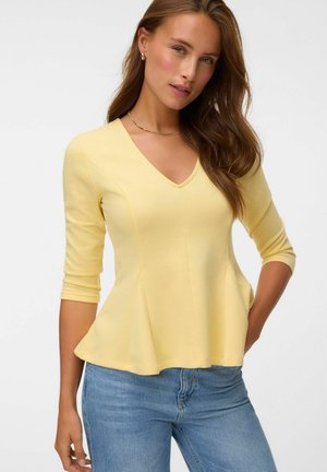 Jeune femme aux longs cheveux bruns porte un haut jaune ajusté à manches trois quarts et un jean bleu, posant devant un fond blanc uni.