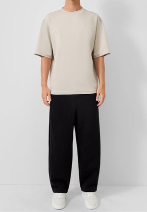 Personne portant un t-shirt beige à manches courtes, un pantalon large noir et des baskets blanches, debout devant un fond gris uni.