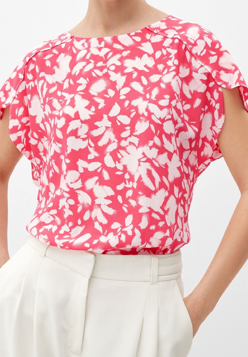 Femme portant un chemisier à manches courtes à motifs floraux roses et blancs, associé à un pantalon blanc taille haute, les mains dans les poches.