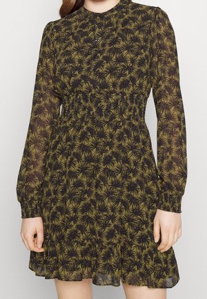 Femme portant une robe à manches longues vert olive avec un motif floral noir épineux, devant boutonné, taille élastique, et ourlet à volants.