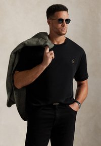 Hombre con camiseta negra y gafas de sol, sosteniendo una chaqueta gris con espiga sobre el hombro, con la mano en el bolsillo de los pantalones negros.