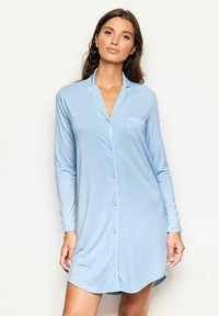 Kvinna iklädd en ljusblå, långärmad, knäppt nattskjorta med vita pipingar och en bröstficka, stående mot en enfärgad ljus bakgrund.