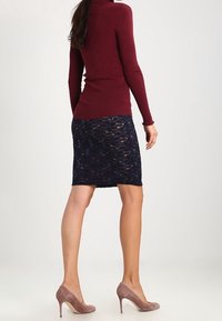 Burgundy anpassad polotröja som matchas med en marinblå spetskjol. Modellen bär taupe högklackade skor, vilket framhäver en elegant silhuett.