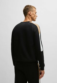 Sweatshirt noir avec un design raglan présentant des accents beiges et blancs sur les épaules. Des lignes nettes et des poignets côtelés créent un look ajusté.