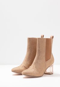 Las botas de tobillo en tono dorado presentan una parte superior de malla texturizada, paneles laterales elásticos, punteras puntiagudas y tacones en bloque transparentes.
