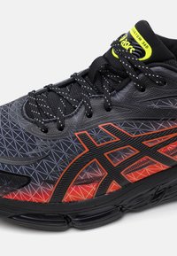 Chaussure de sport noire avec un motif géométrique, des accents rouges et orange, un matériau texturé et une semelle rembourrée avec des chambres à air visibles.