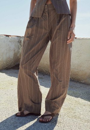 Persona que lleva puestos pantalones sueltos marrón con rayas, sandalias marrones, de pie al aire libre sobre una superficie de hormigón texturizado cerca de un muro bajo y envejecido.