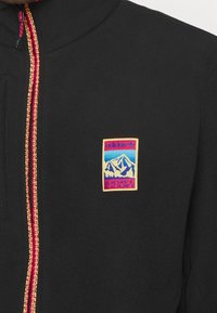 Černá fleecová bunda s zipovým uzávěrem, zdobená vícolorovými detaily a nášivkou s horským motivem v modré a žluté barvě na hrudi.