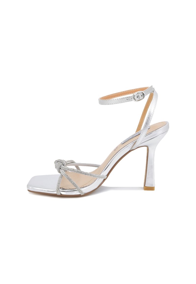 Sandalo con tacco argento, punta quadrata, cinturino alla caviglia e dettaglio a nodo decorativo. Materiale lucido con accenti testurizzati e tacco sottile.
