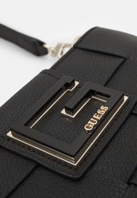 Czarna torebka ze skóry o fakturze, z błyszczącym czarnym logo i złotymi akcentami, prostokątny kształt z paskiem do noszenia.