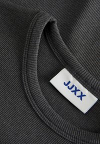 Harmaa rypytetty kangas, jossa on pyöreä pääntie ja valkoinen etiketti, jossa lukee "JUXX" sinisellä. Lähikuvassa korostuu kankaan rypytys.