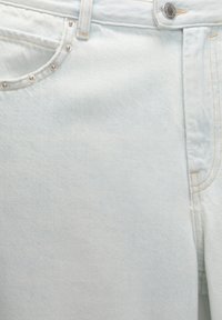 Jean en denim clair présentant une couleur bleue délavée, un design à cinq poches, des clous décoratifs argentés le long de la poche avant, et des coutures contrastantes.