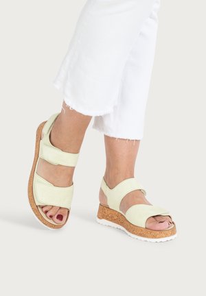 Füße in hellgrünen Keilsandalen mit Korksohlen und weißen Außensohlen, kombiniert mit weißen kurzen Hosen mit ausgefransten Säumen.