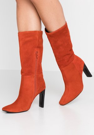 Jambes portant des bottes à talons en suède orange montant jusqu'aux genoux, avec des fermetures éclair sur le côté et des talons carrés noirs sur fond blanc.