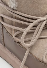 Bota marrón claro con detalles de cordones, textura suave y suela sintética. Cuenta con acentos marrones y un parche de piel cerca de la parte superior para mayor detalle.