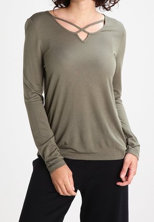 Frau in einem olivgrünen Langarmshirt mit gekreuzten Trägern am Ausschnitt und schwarzer Hose, stehend mit einer Hand nahe der Tasche.