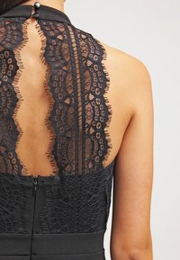 Černé šaty s krajkovým detailem na zádech, s vysokým výstřihem a viditelným zipem. Krajka má květinový vzor a třepící se okraje.