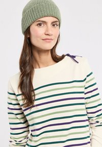 Femme aux longs cheveux bruns portant un bonnet en tricot vert clair et un pull crème avec des rayures horizontales vertes, violettes et bleu foncé.