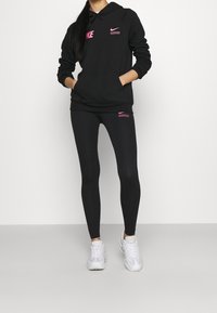 Svart hoodie med rosa Nike-logotyp på bröstet och fickan, ihop med svarta leggings som har en liten rosa Nike-logotyp. Vita sneakers.