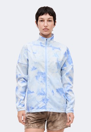 Άτομο που φοράει ένα ανοιχτό μπλε και άσπρο μοτίβο μπουφάν με φερμουάρ και λογότυπο στο στήθος και καφέ σορτς, στέκεται μπροστά σε απλό φόντο.