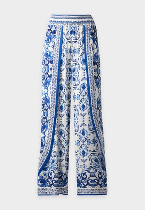 Femme portant un chemisier à manches courtes blanc et bleu à motifs et une jupe longue assortie, associée à des sandales blanches à talons hauts, debout.