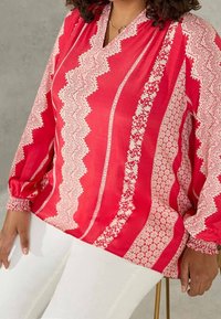 Rote Bluse mit weißen geometrischen Mustern, V-Ausschnitt, langen Puffärmeln und elastischen Bündchen, kombiniert mit weißen Hosen.