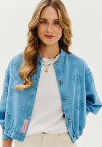 Světlomodrá vyšívaná bunda s kulatým výstřihem, zapínáním na knoflíky a elastickými manžetami, nošená přes bílý top a béžové kalhoty.