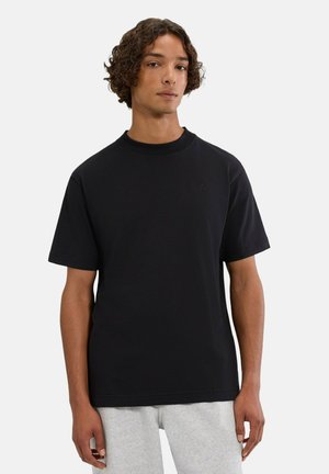 Junger Mann mit lockigem Haar trägt ein schlichtes schwarzes kurzärmeliges T-Shirt und hellgraue Hose und steht vor einem weißen Hintergrund.