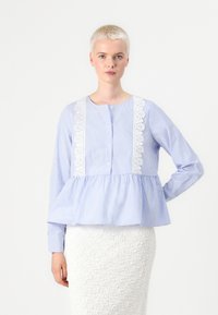 Modra in bela prugasta bluzica s peplum krojem in dolgimi rokavi, okroglo izrezom in čipkastimi detajli spredaj. Zapenjanje na gumbe.