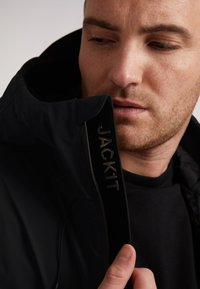 Giacca nera con colletto alto, dotata di chiusura con zip e branding "JACK1T". Tessuto testurizzato e elementi di design discreti visibili.