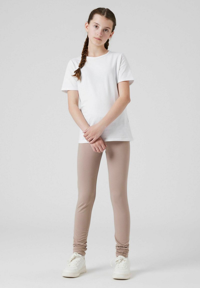 Witte katoenen T-shirt gecombineerd met lichtbeige leggings. De leggings hebben gerimpelde details bij de enkels. Model draagt witte sneakers.