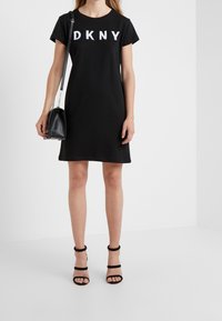 Černé bavlněné šaty s krátkým rukávem, s bílým nápisem "DKNY". Doplněno černými sandály na vysokém podpatku se pásky a malou černou kabelkou.