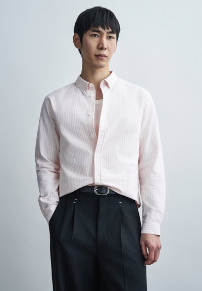 Chemise rose clair à rayures avec col étalé, associée à un pantalon foncé doté d'une boucle argentée et de coutures apparentes.