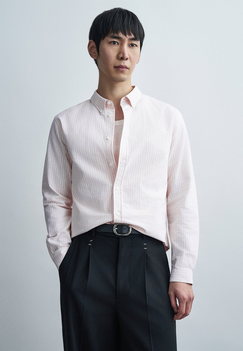 Ροζ ριγέ πουκάμισο με κουμπιά και γιακά spread, σε συνδυασμό με σκούρο παντελόνι που έχει ασημένια αγκράφα και εμφανείς ραφές.