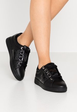 Czarne sneakersy niskie z tłoczonymi panelami i czarnymi sznurówkami noszone na stopach na tle białym.