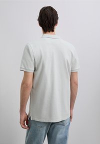 Lichtgrijs poloshirt met een gestructureerde afwerking, korte mouwen en zijgaten. Draagt bij een lichtblauwe spijkerbroek. Minimalistisch ontwerp.